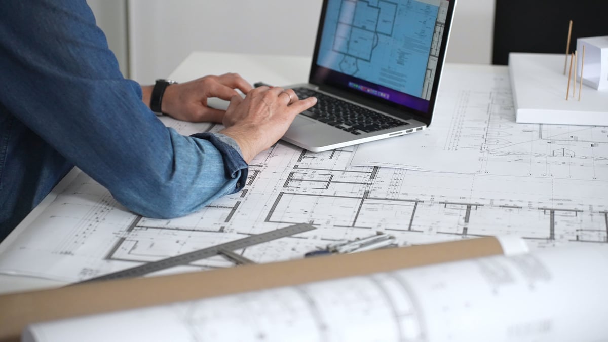 Architect drawing building blueprints on a white desk with laptop displaying floor plans